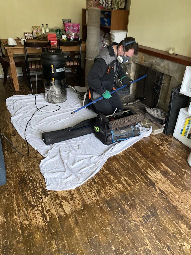Professional chimney sweep using HEPA vacuum system with protective dust sheets laid around fireplace during cleaning process in Cambridge home